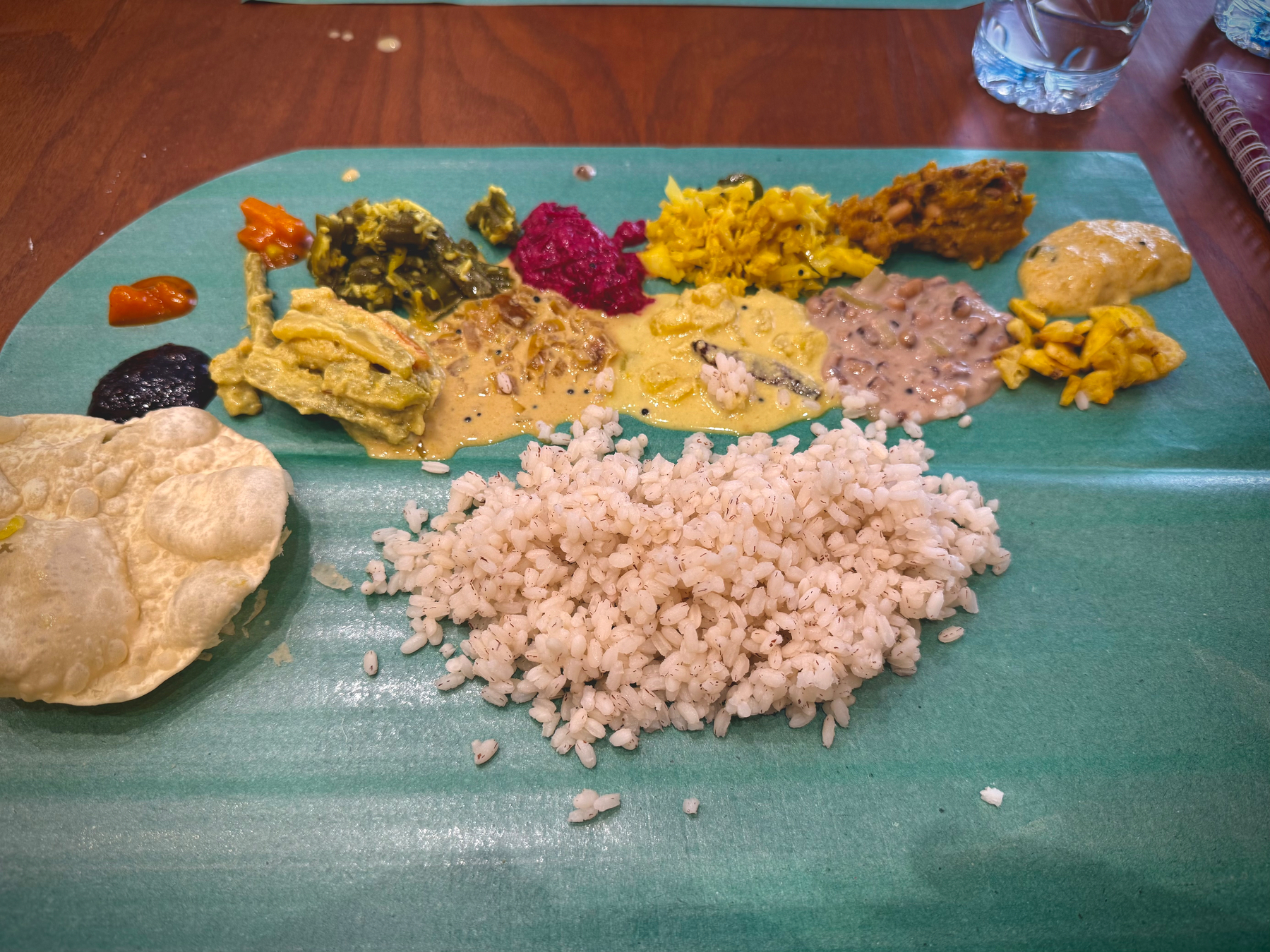 Auto-generated description: A traditional Sadya, a South Indian meal, is served on a banana leaf, featuring a variety of chutneys, curries, papad, and steaming rice.