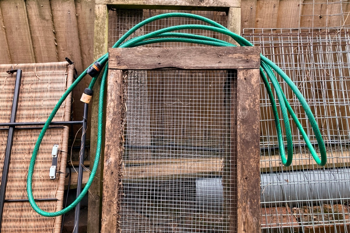 Photo of a green hosepipe draped over a load of wooden panels leaning against a wooden fence.
