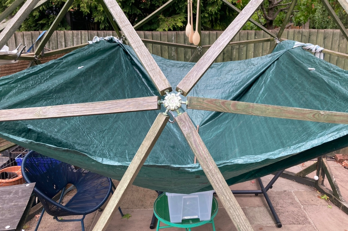 A view into the geodesic dome. A joint of the dome is in the middle of the frame with six poles coming out of it. Behind is a green gazebo tent covering strung between four joints. Under the middle is a large plastic container on a small table.