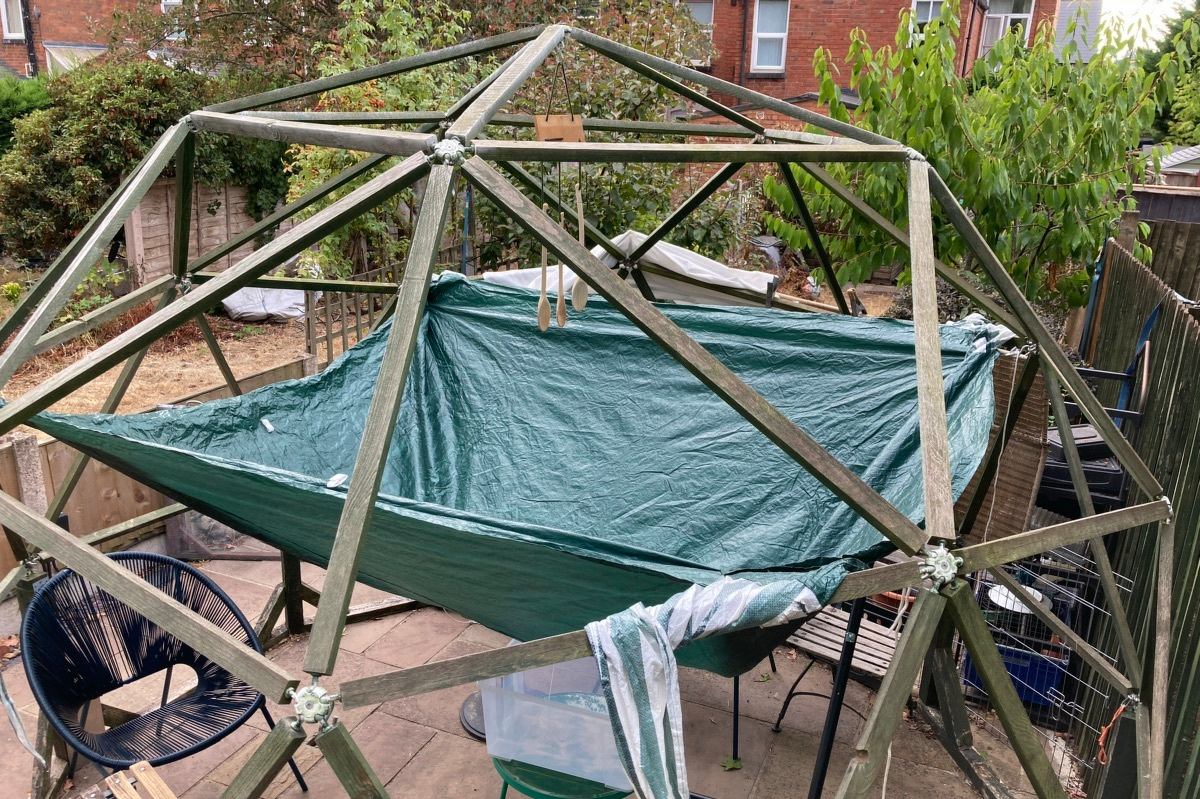 A view of the geodesic dome from above and to the side. A green gazebo tent is hung from four joints of the dome. Under the middle is a large plastic container on a small table.