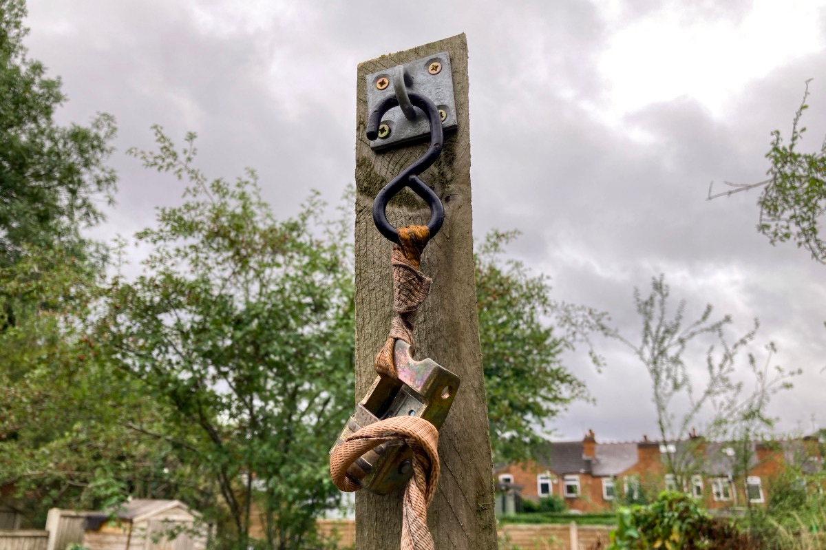 Photo of a wooden post reaching into the overcast sky. It has a hoop screwed into the top and a ratchet strap is hanging from it. The strap is old and worn and twisted.