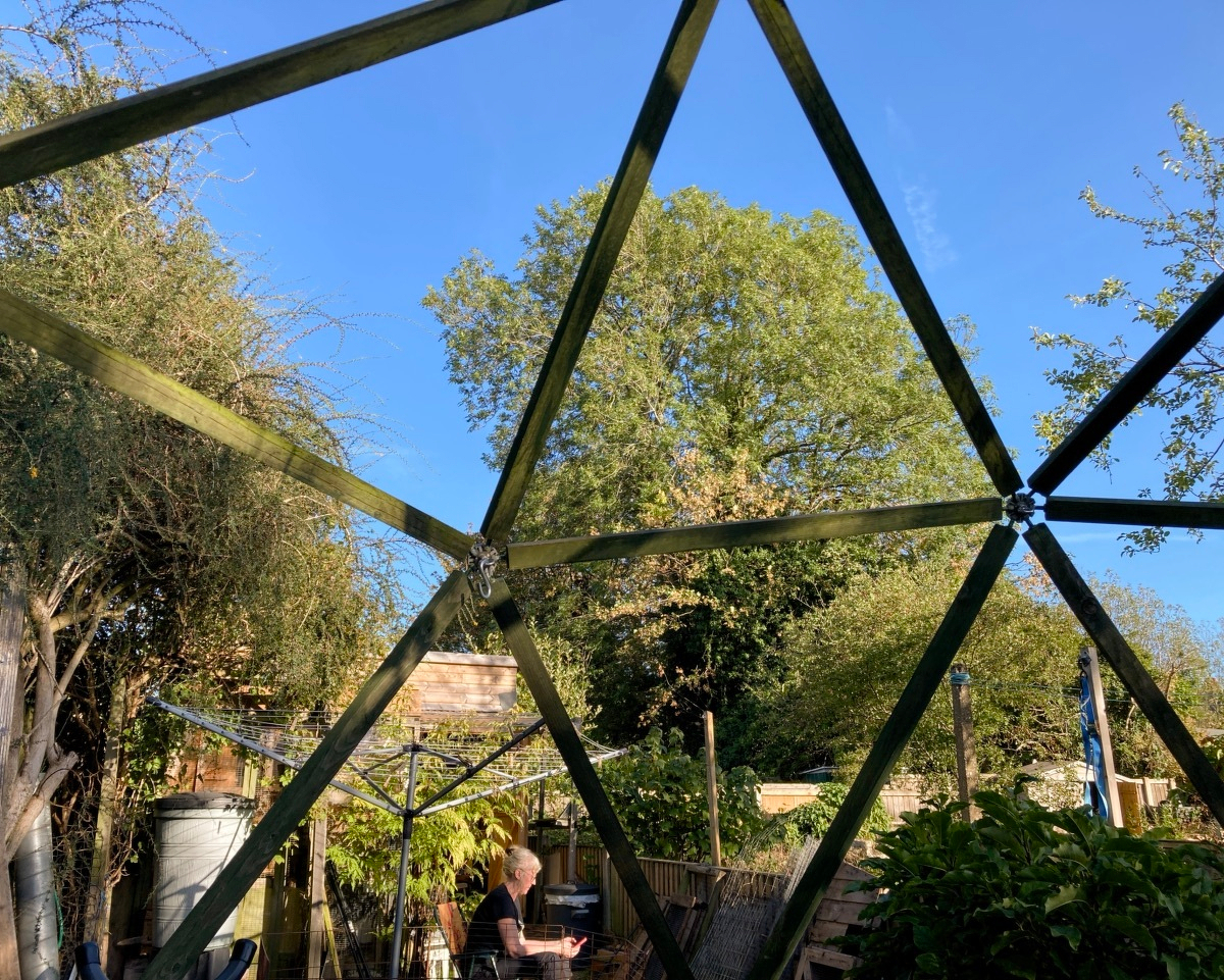 View through the beams of the geodesic dome in our back garden. The blue sky and trees are in the background. A woman is sitting in the lower foreground. A shed and various garden detritus are around her.