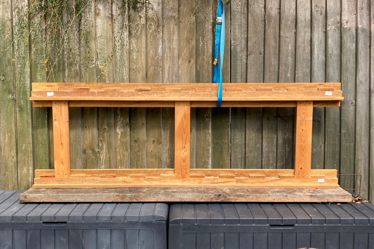 A wooden frame made up of random pieces of wood is resting on some plastic storage chests against a green wooden fence. A blue strap is holding it in place somehow.