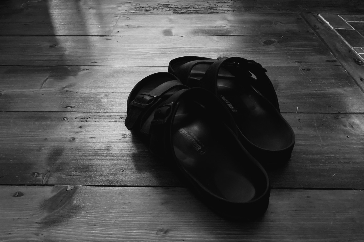 A black and white photo of a pair of black sandals on a wooden floor with light coming from the background.