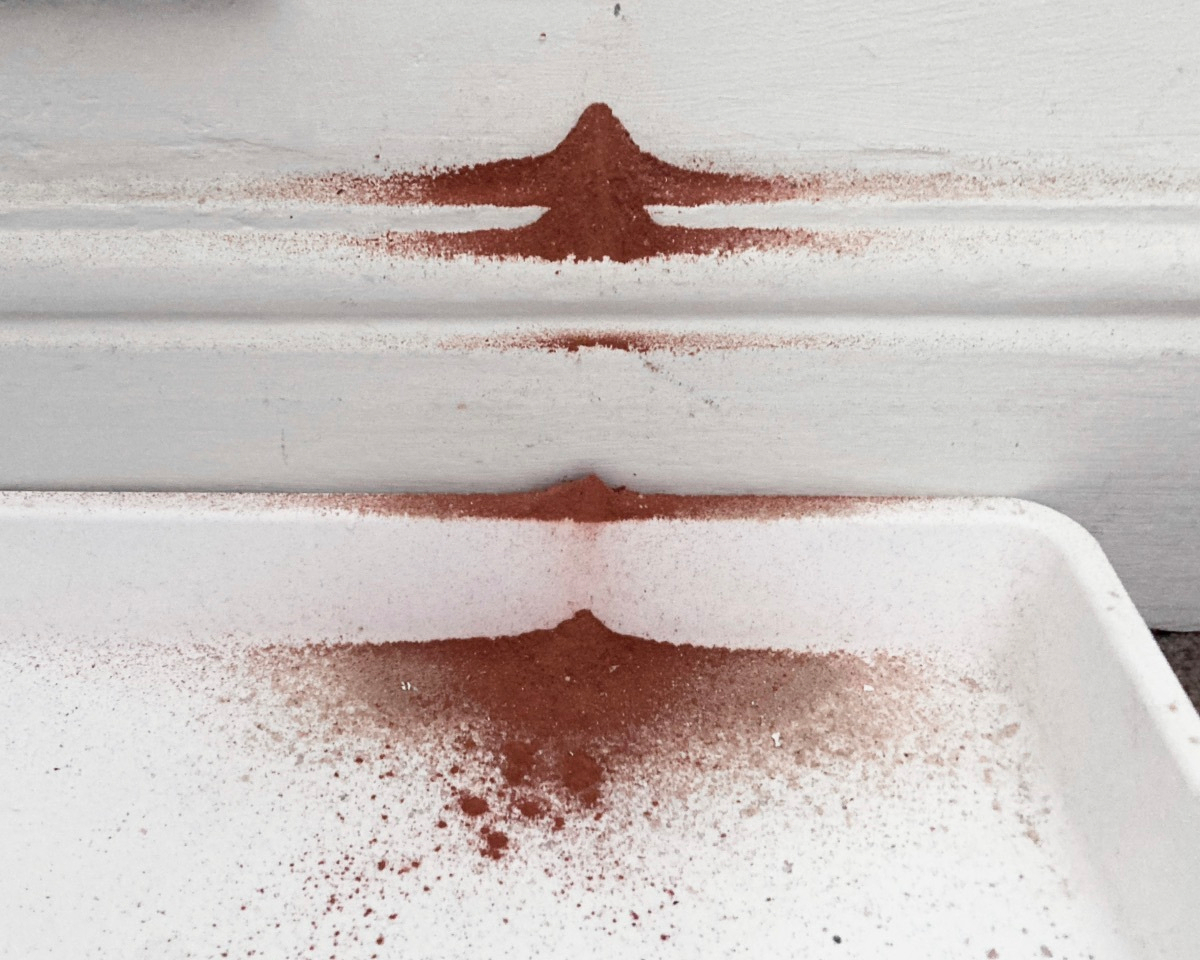 Brick dust from drilling holes in the wall above has collected on the skirting board and in a plastic container on the ground. It has settled in small mounds on each ridge in its way.