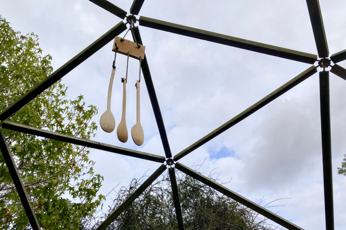 Three wooden spoons are hanging like a wind-chime from a joint on a geodesic dome. The sky is visible through the dome and is overcast.
