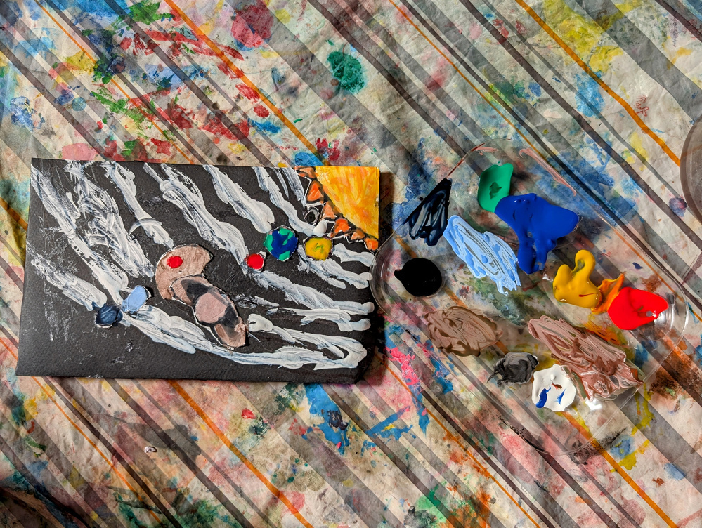 A small abstract painting on a colorful, splattered surface is surrounded by various blobs of bright paint and mixing tools.