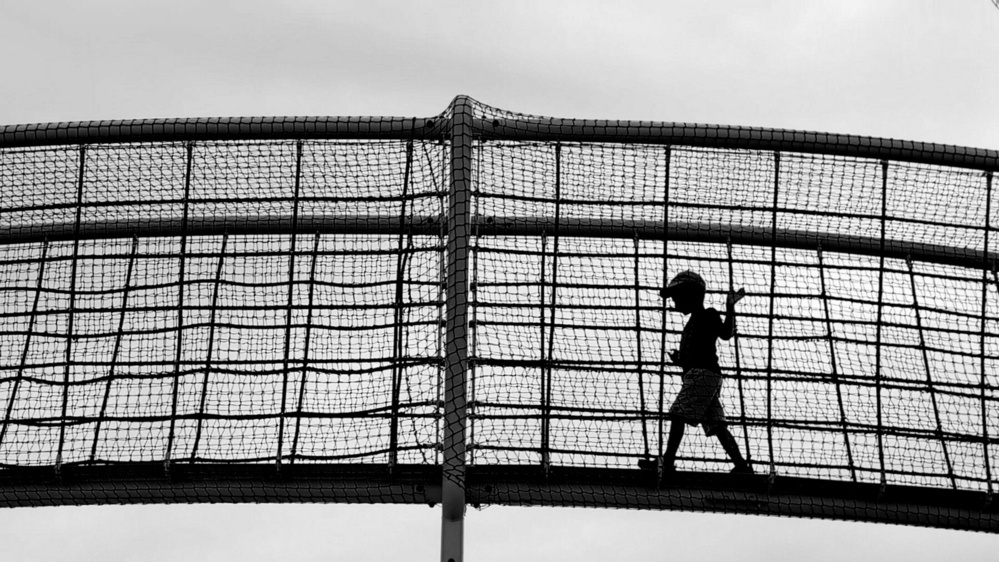 A child walks across a mesh-covered bridge silhouetted against the sky.