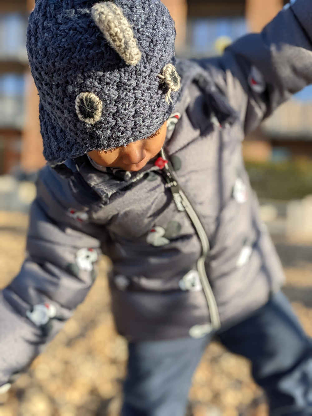 A young child bundled in a grey jacket and a knitted blue hat with playful animal features is outdoors, stretching their arms to the sides.