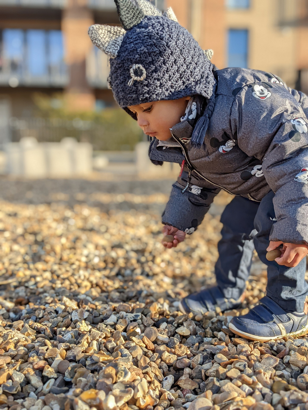 A young child wearing a dinosaur-themed knit hat and a jacket with Mickey Mouse prints is bent down and closely examining the gravel on the ground.
