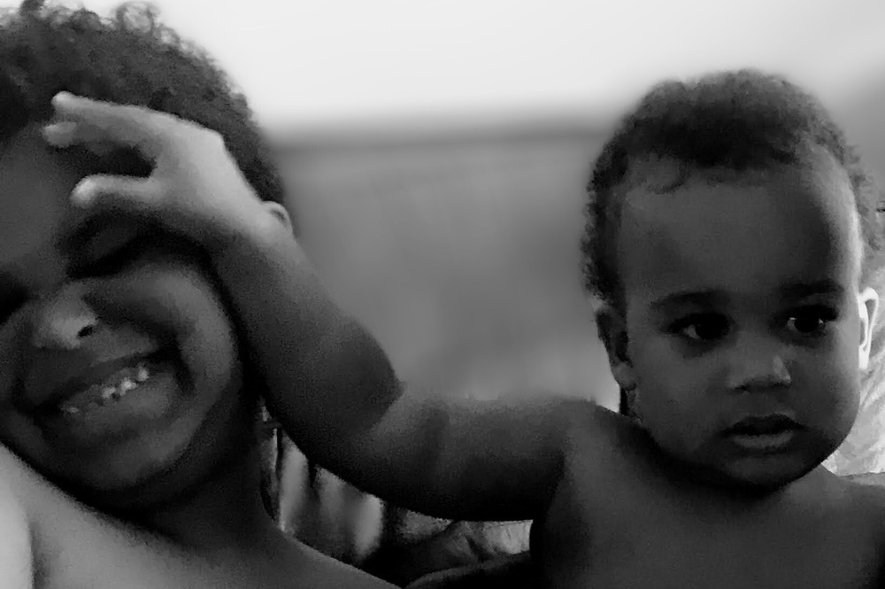Two young children are playfully interacting, with one child smiling while the other reaches out to touch their face.