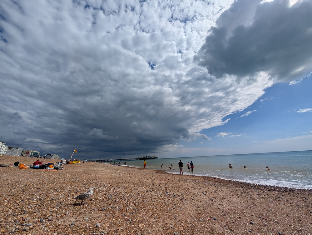 A pebble beach with people swimming and relaxing under a dramatic sky where dark clouds are meeting clearer blue skies.