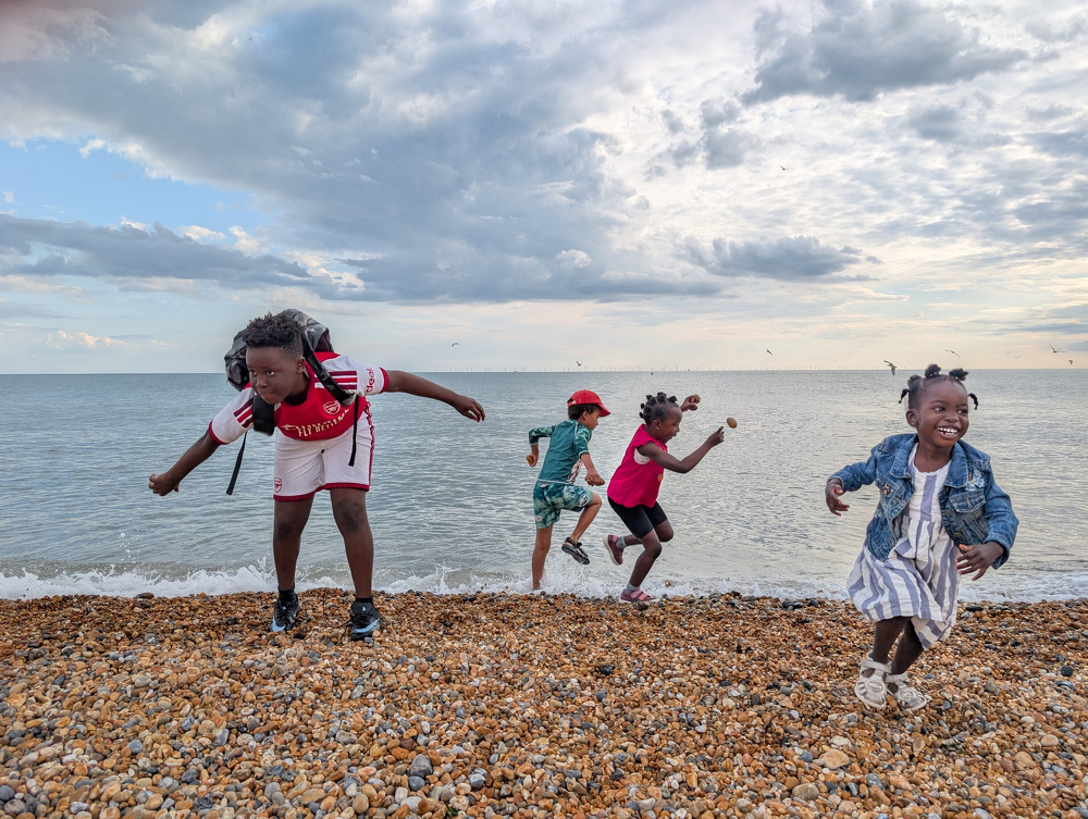 Four children are joyfully playing and jumping on a pebble beach by the sea under a cloudy sky.