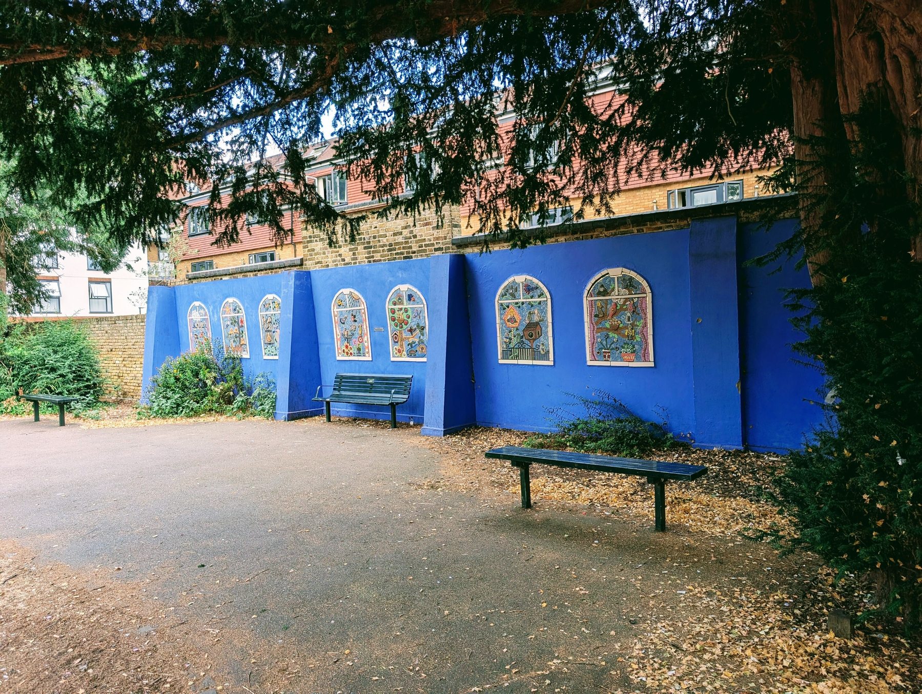 Colourful mosaic panels on a blue wall depict the old park aviary which once stood on this spot in front of a new housing block with park benches in front and tree cover overhanging the frame.