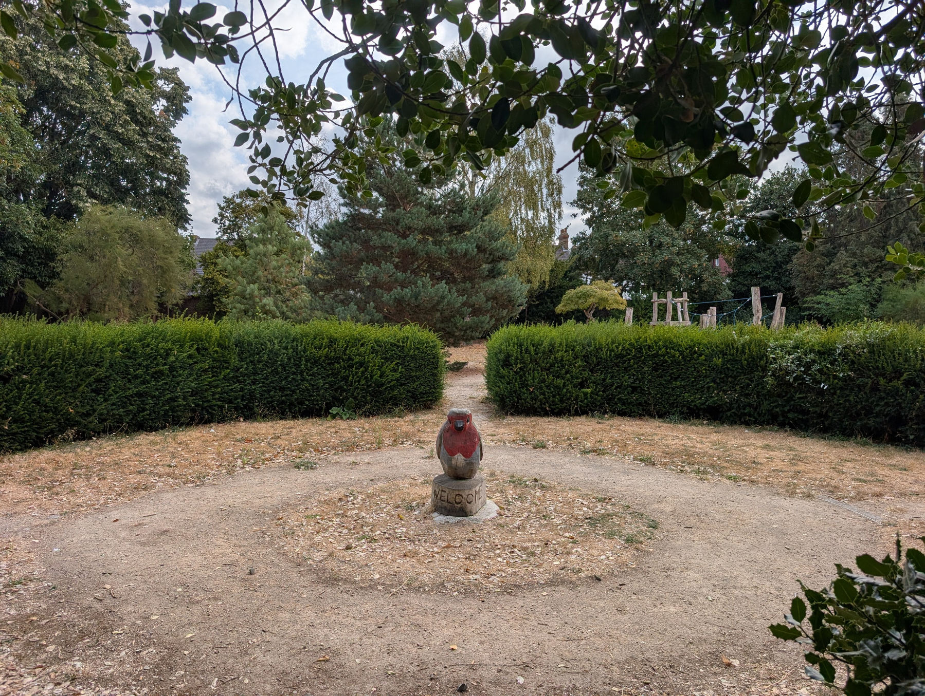 A giant wood-carved robin with a painted red breast stands atop a circular wooden plinth with WELCOME etched into it amid a circular sunburnt garden and path surrounded by green hedgerows and mature trees in the background.