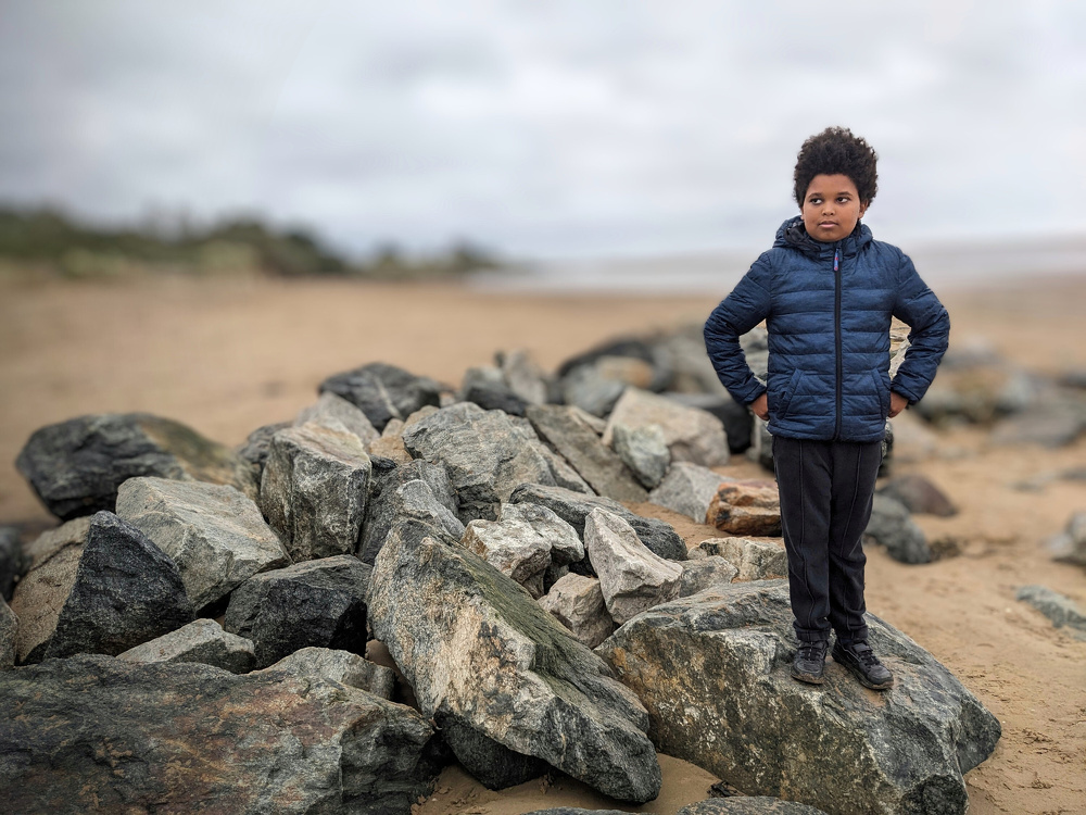 A young boy wearing a blue jacket stands on a large rock amidst a pile of rocks on a cloudy beach.