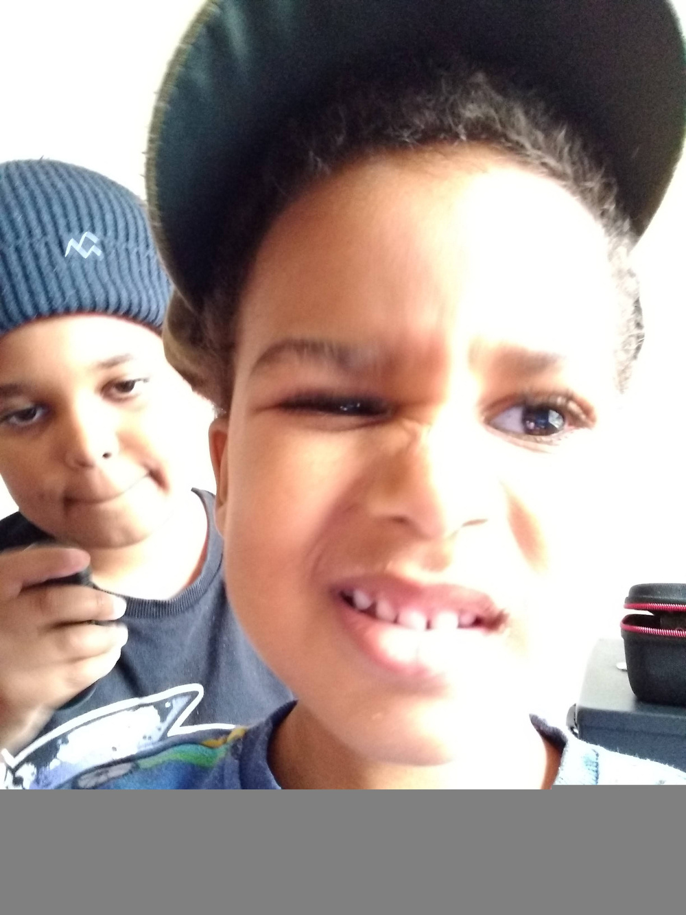 Two young boys are posing closely to the camera, with one boy in the foreground making a scrunched-up facial expression and the other boy in the background wearing a knit hat.