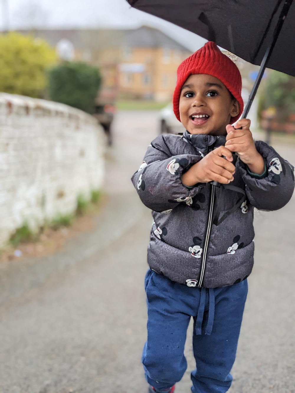 A young child wearing a red beanie and a Mickey Mouse jacket is happily holding a black umbrella while standing on a pathway.