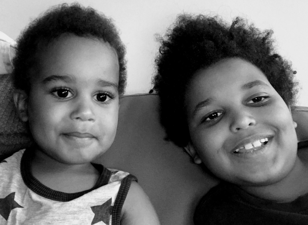 Two young children with curly hair are sitting closely together, one smiling and the other with a neutral expression.