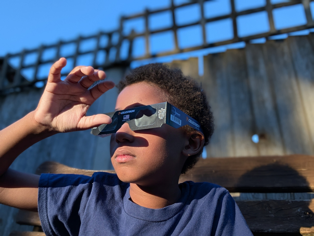 A young boy is looking up at the sky while wearing solar eclipse glasses and holding them with one hand.