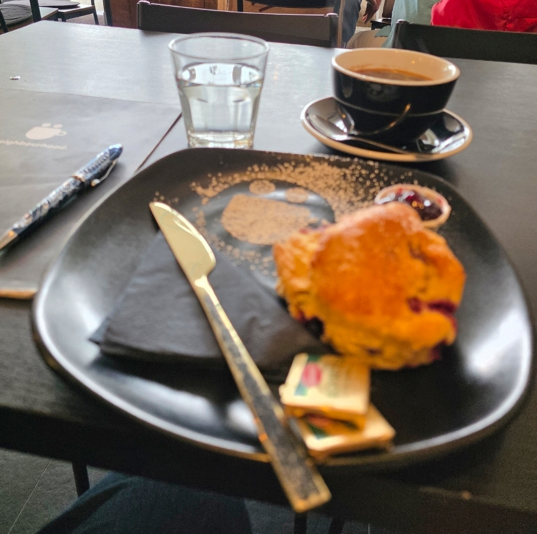  A scone with Kerrygold butter and jam, a cup of coffee, and a glass of water are arranged on a table along with a Rhodia notebook and a Snake pen.