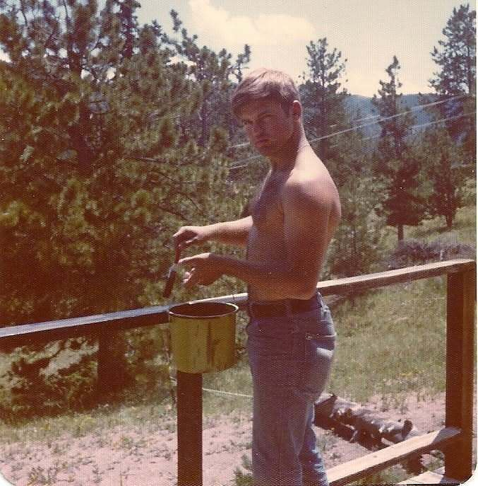 Pat Petersen the younger, on a wooden deck in a forested area, holding a paintbrush and painting a railing.