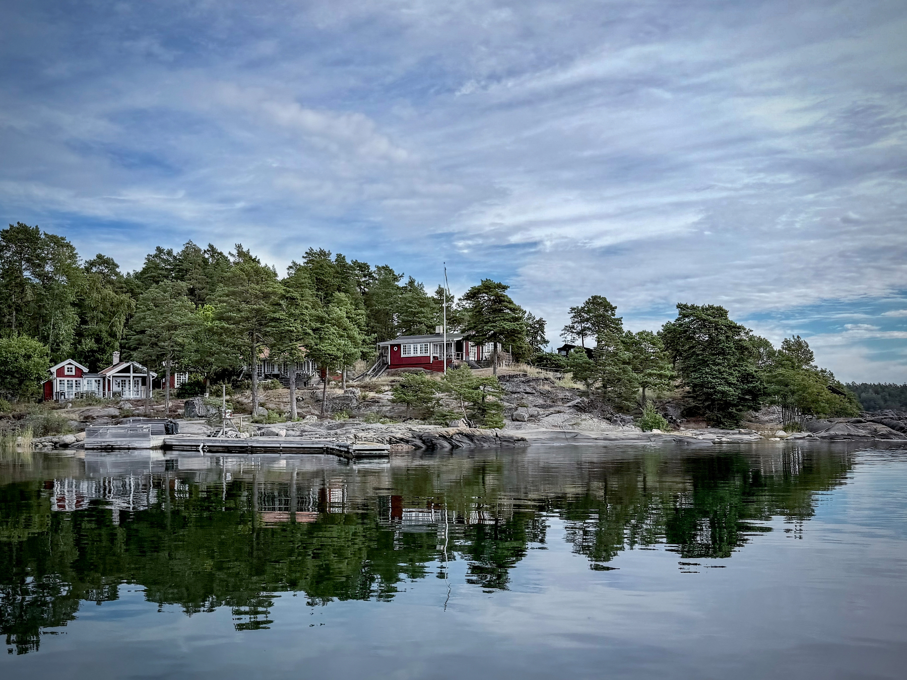 En lugn skärgårdsmiljö med hus, klippor och träd speglade i det stilla vattnet under en molnig himmel.