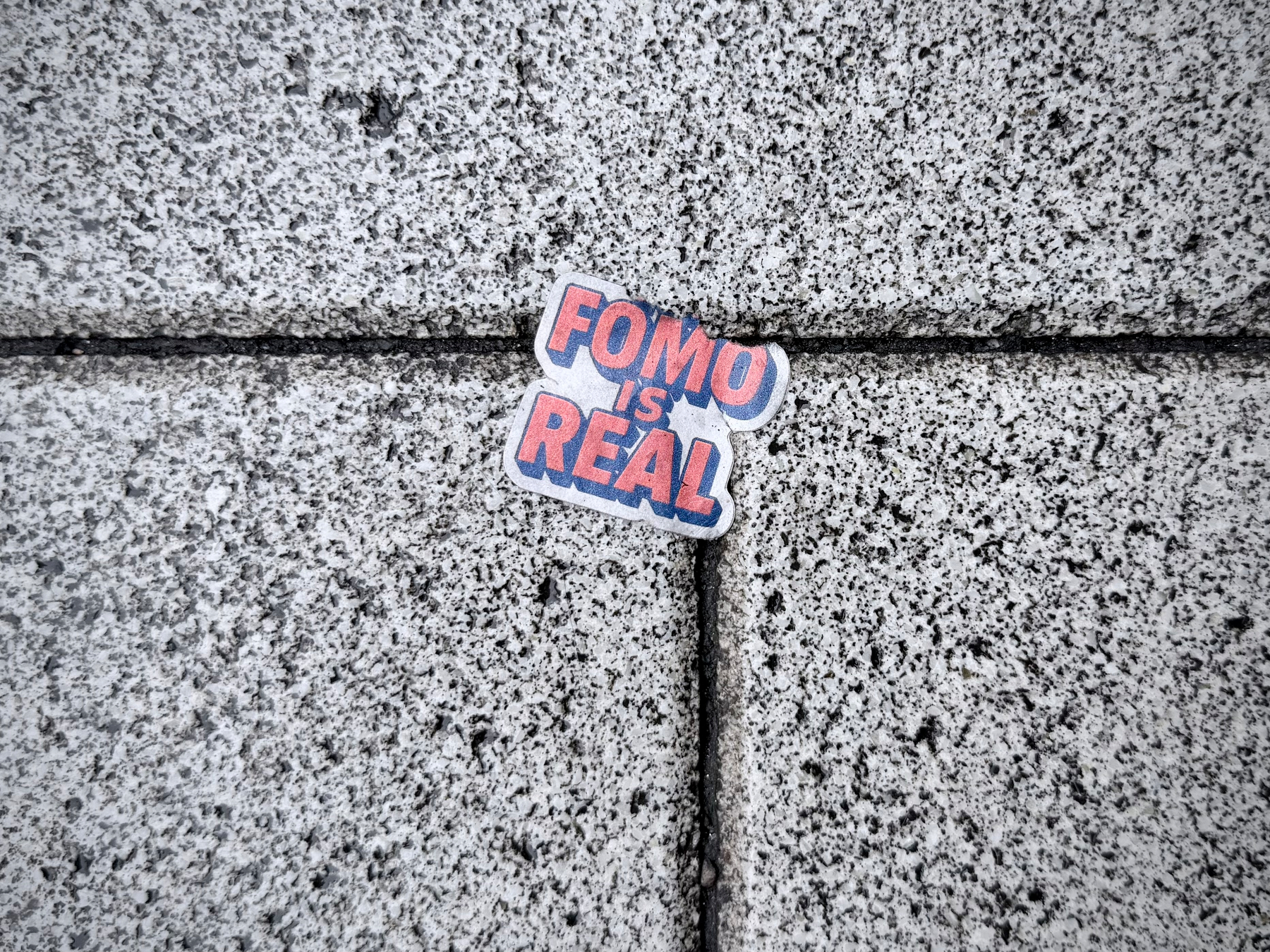 A sticker with the words FOMO IS REAL is placed on a textured gray stone surface at the intersection of four tiles.