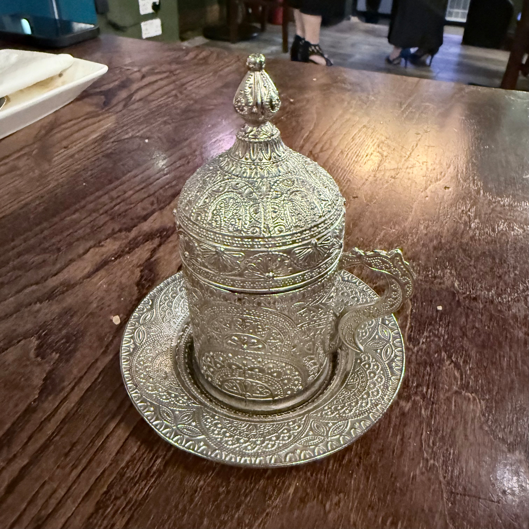 An intricately designed, ornate metal cup with a matching lid and saucer is placed on a wooden table.