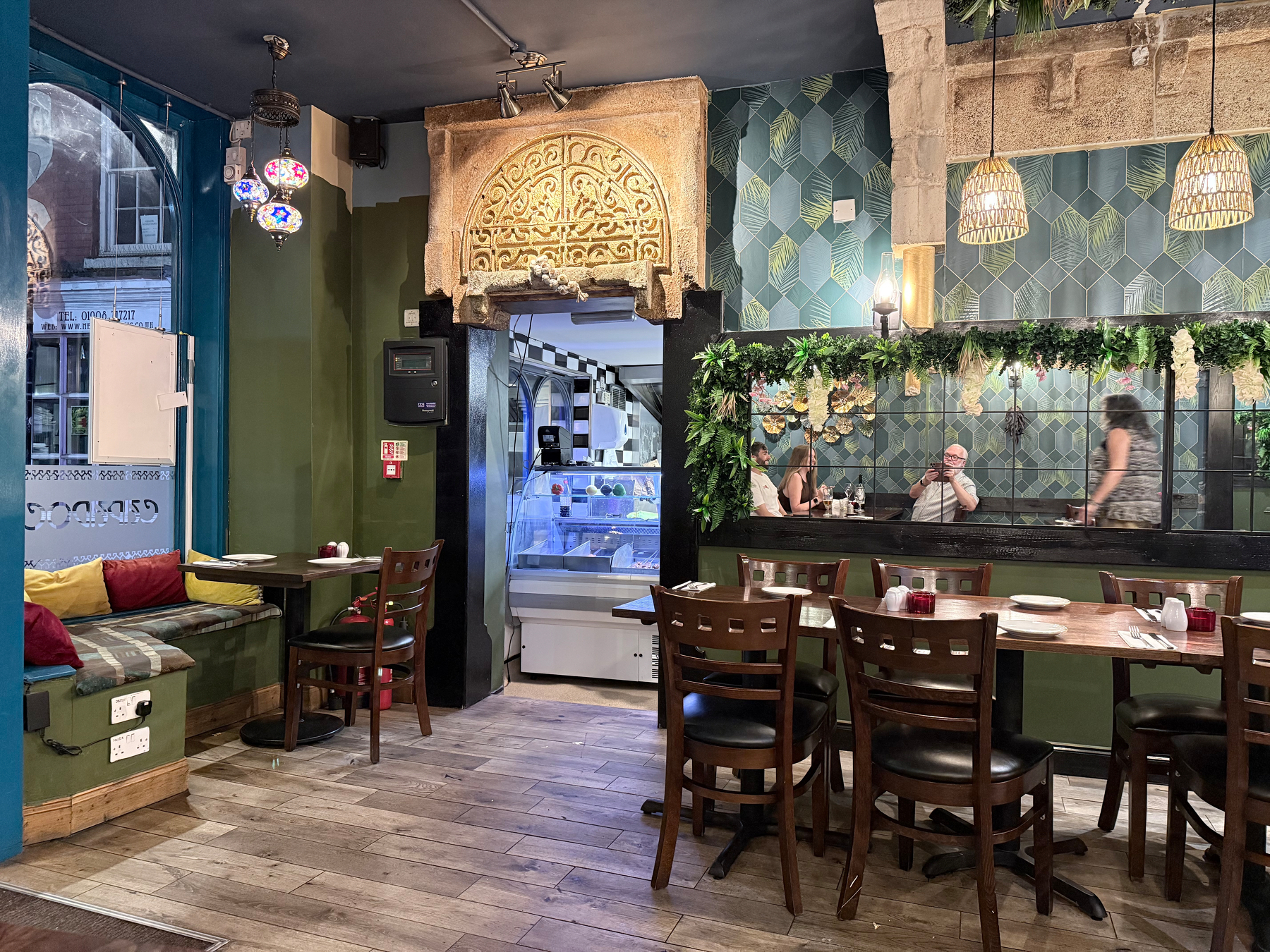 A cozy restaurant interior features wooden tables and chairs, a cushioned seating area by the window, decorative hanging lights, and a green-tiled wall with floral accents.